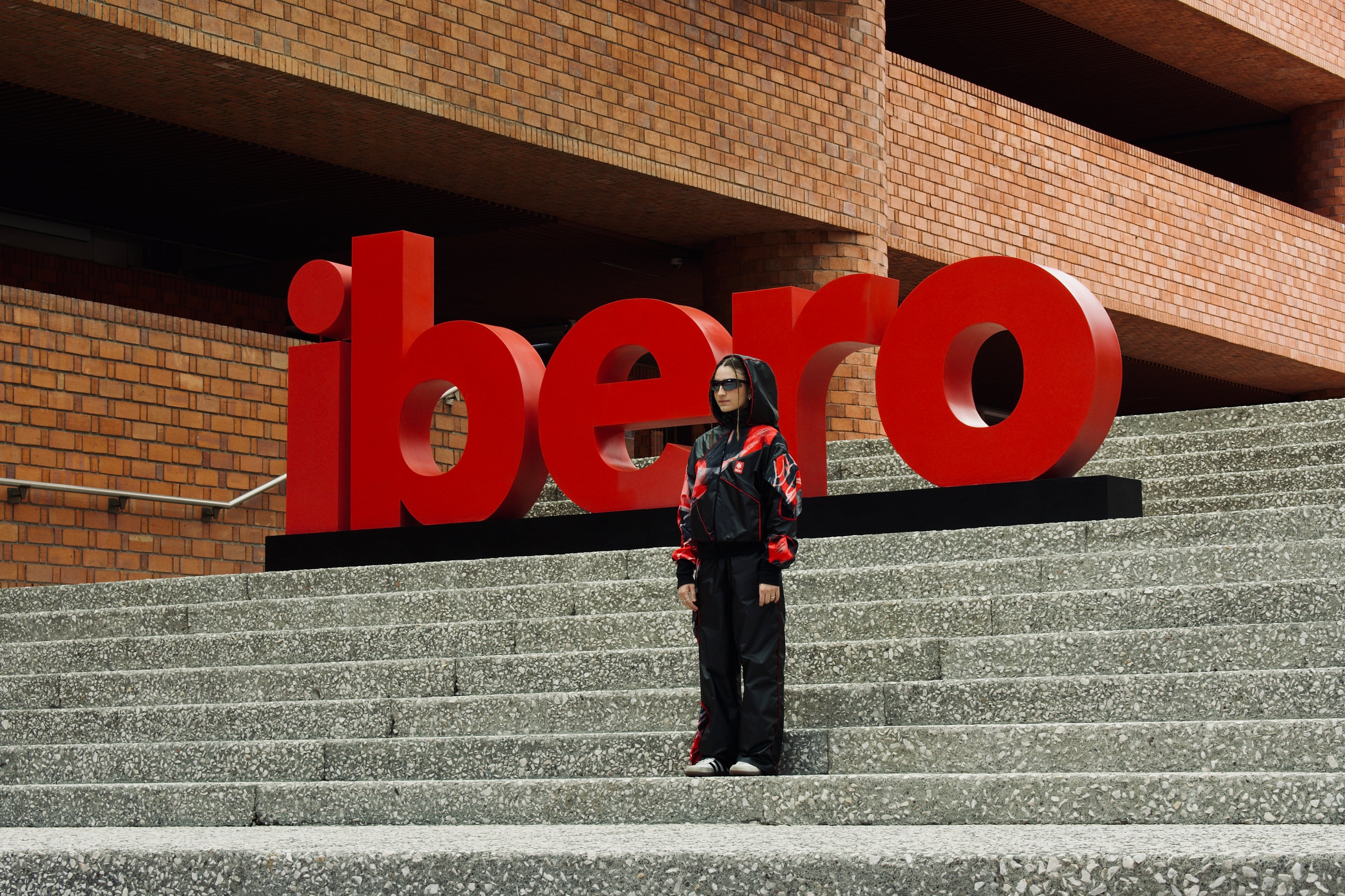 Modelo con el uniforme deportivo negro y rojo frente a la escultura tipográfica “ibero”, en la escalinata de la universidad.
