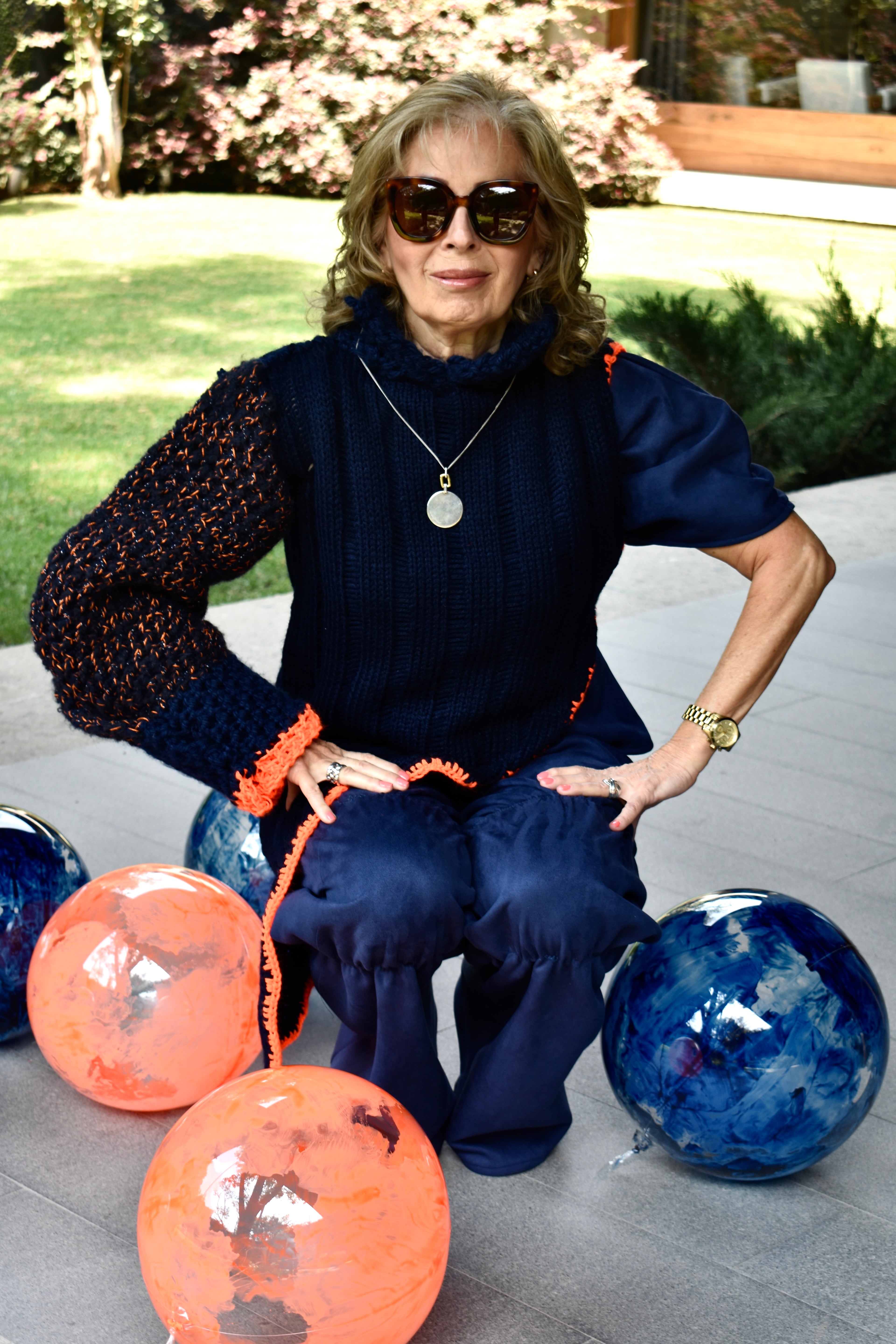 Mujer de la tercera edad sentada en un jardín, vistiendo un suéter tejido azul marino con mangas en punto naranja de la colección Korel, rodeada de esferas de vidrio.
